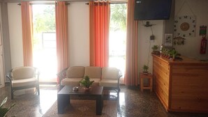 Una sala de estar con sofá, mesa de centro y televisor. Hay cortinas, un reloj y una mesita con plantas.