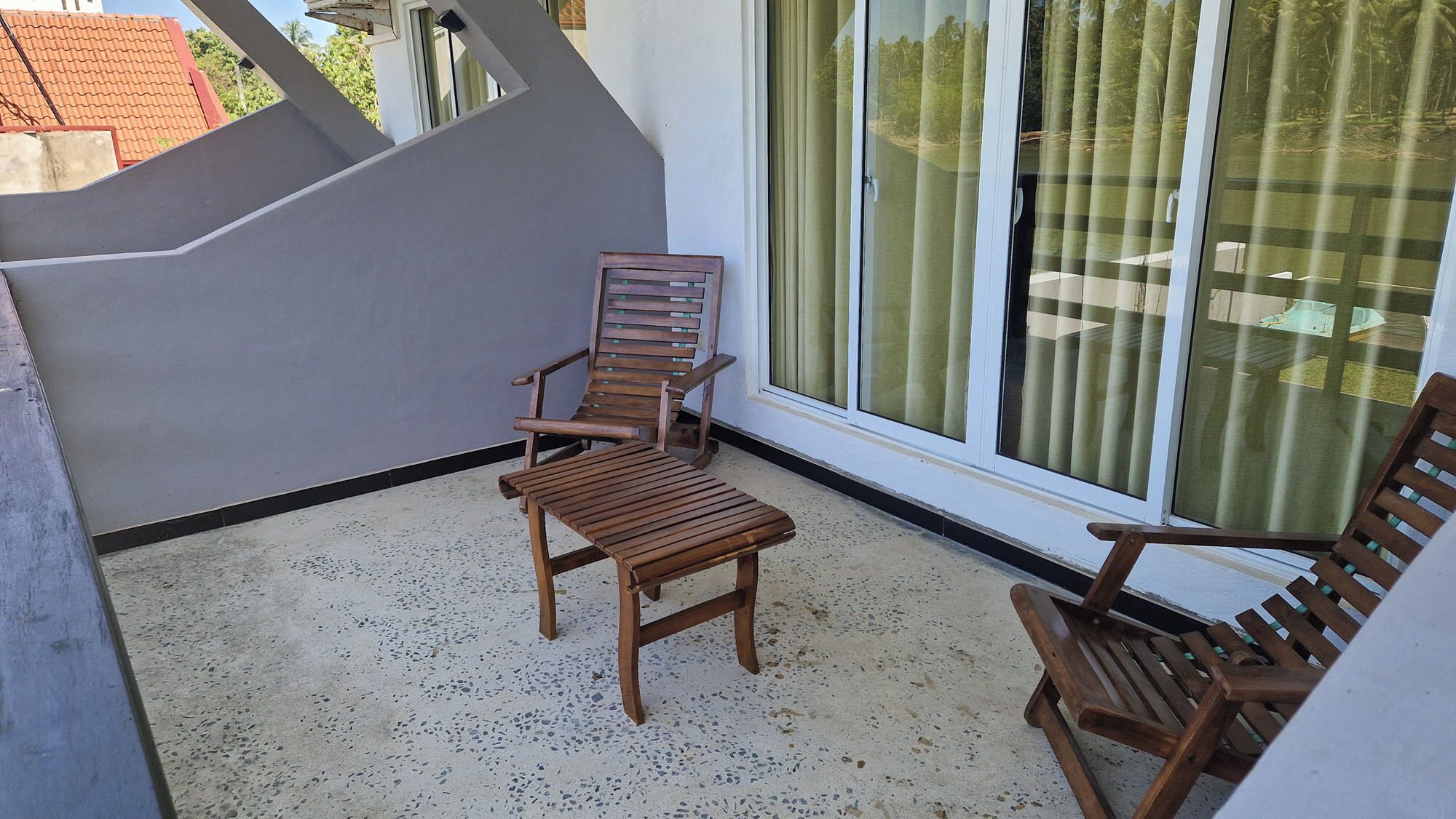 A balcony with two wooden chairs and a small table, a glass door with curtains, and a view of a residential area.