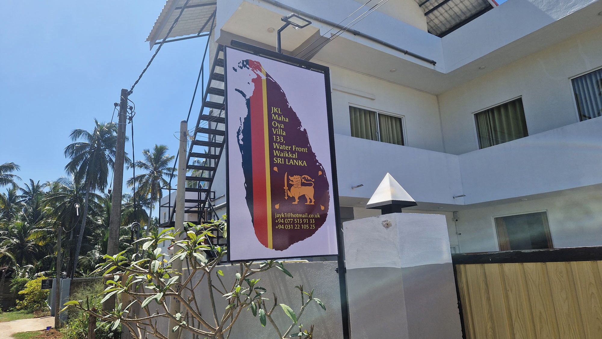 A building with a signboard advertising JKL Maha Oya Villa, including contact information and location details.