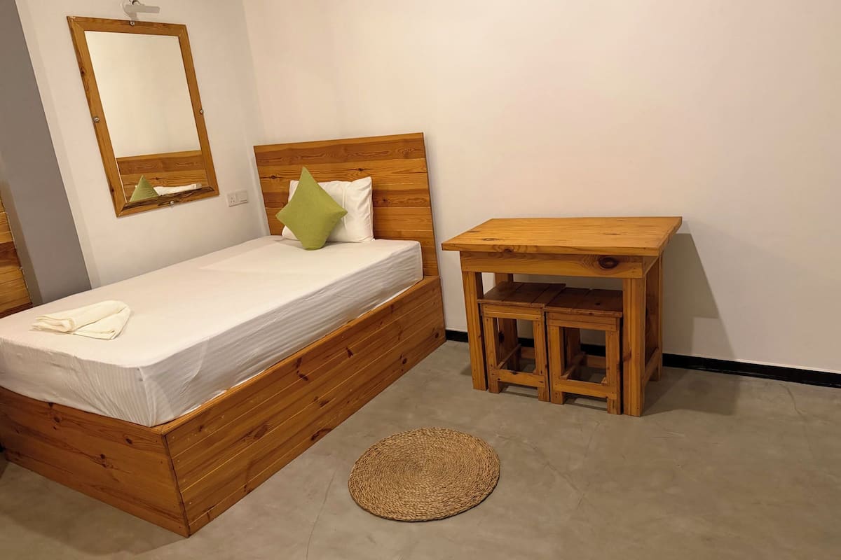A wooden bed with a mattress and pillows, a small wooden table, a round woven mat, and a mirror on the wall.