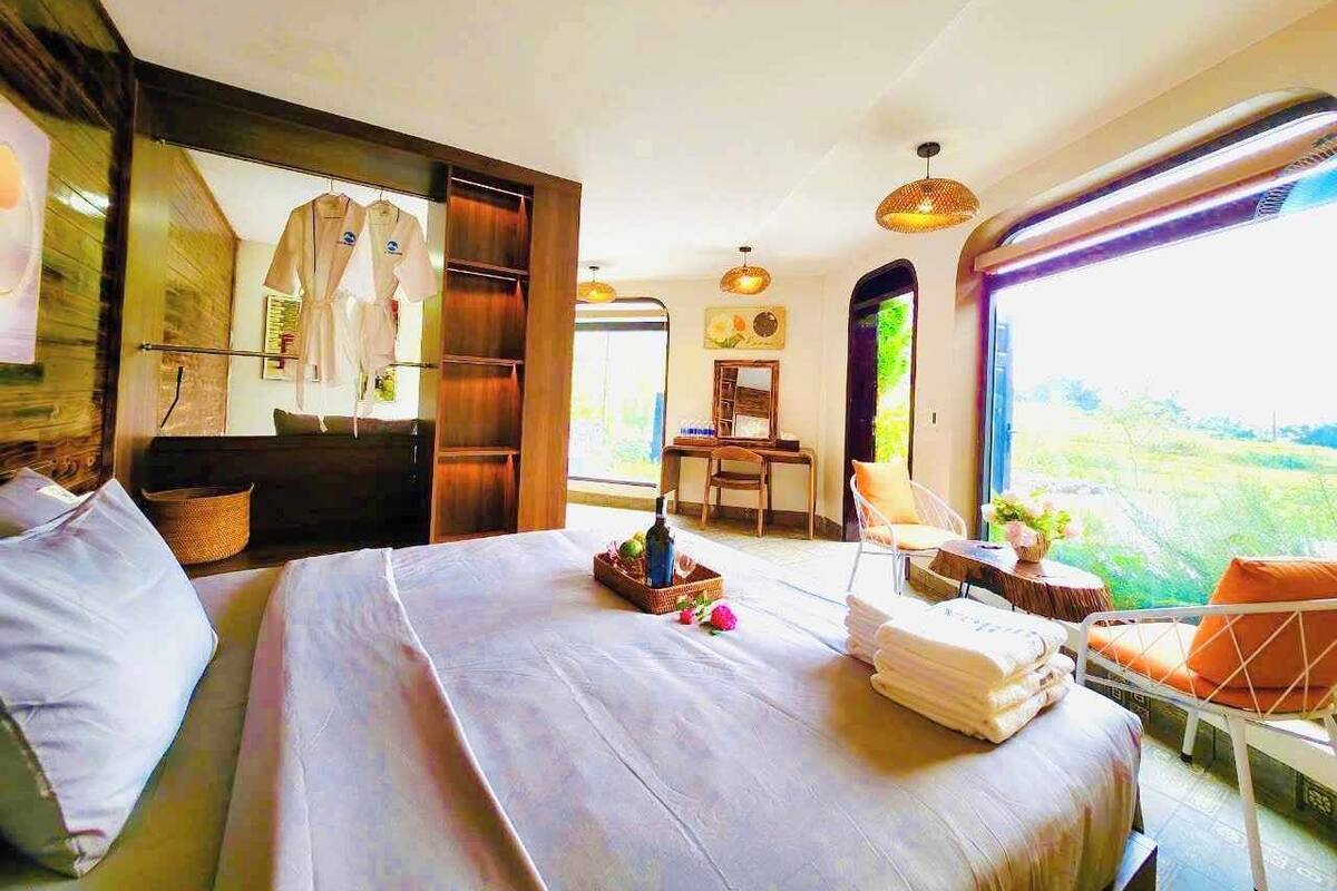 A spacious bedroom with a large bed, a wooden wardrobe, a chair, and a view of greenery outside.