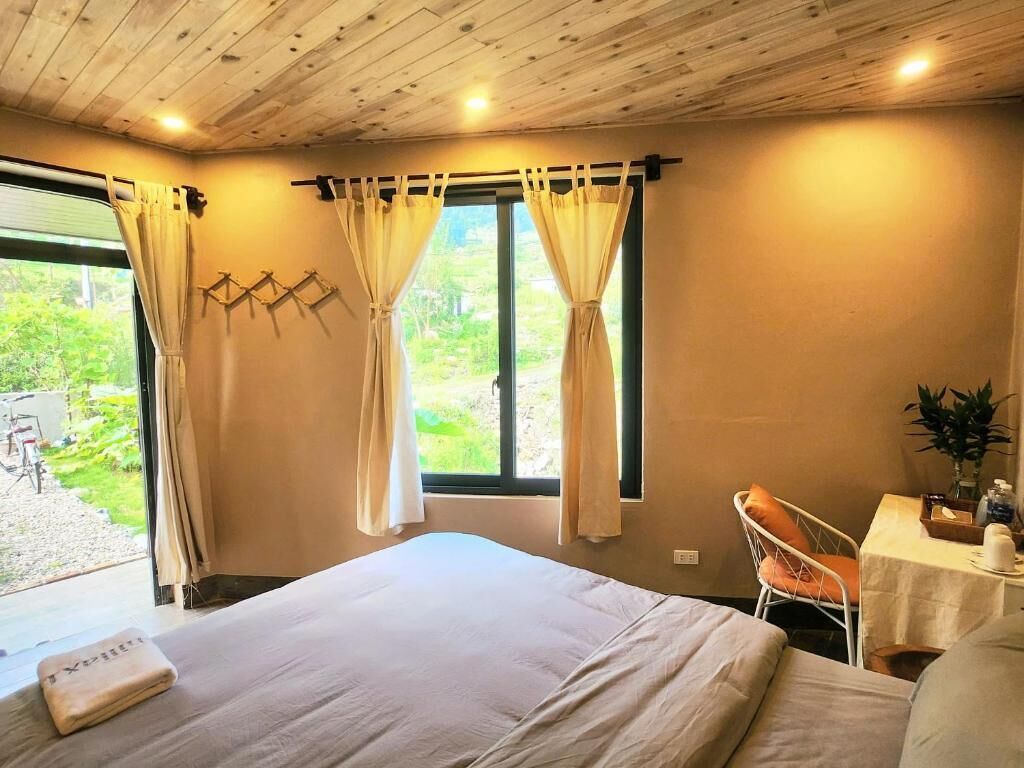 A bedroom with a large bed, two windows with curtains, a small table with a plant, and a chair.