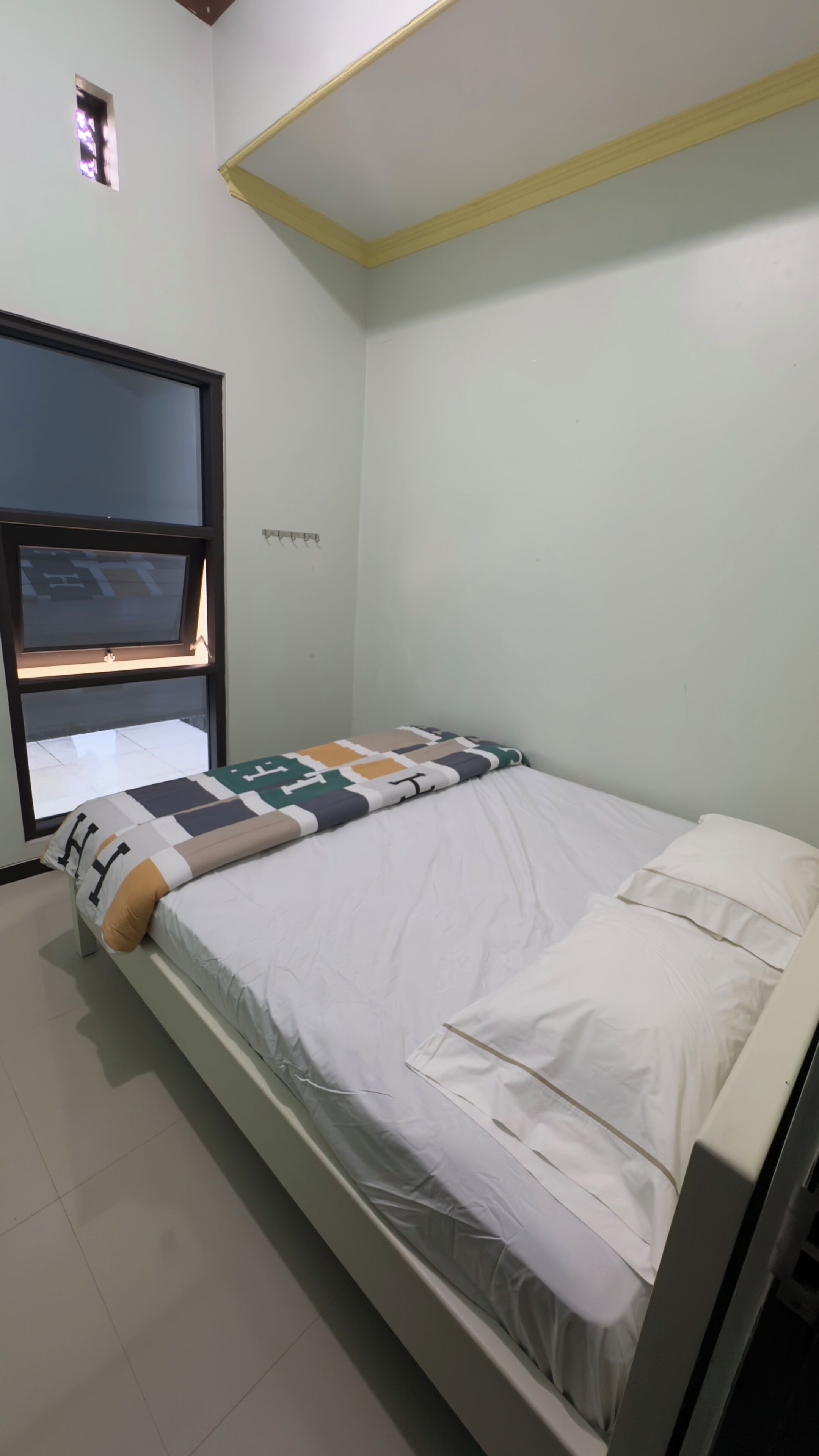 A single-bed room with a window, a bed with a patterned blanket, and a small window above the bed.