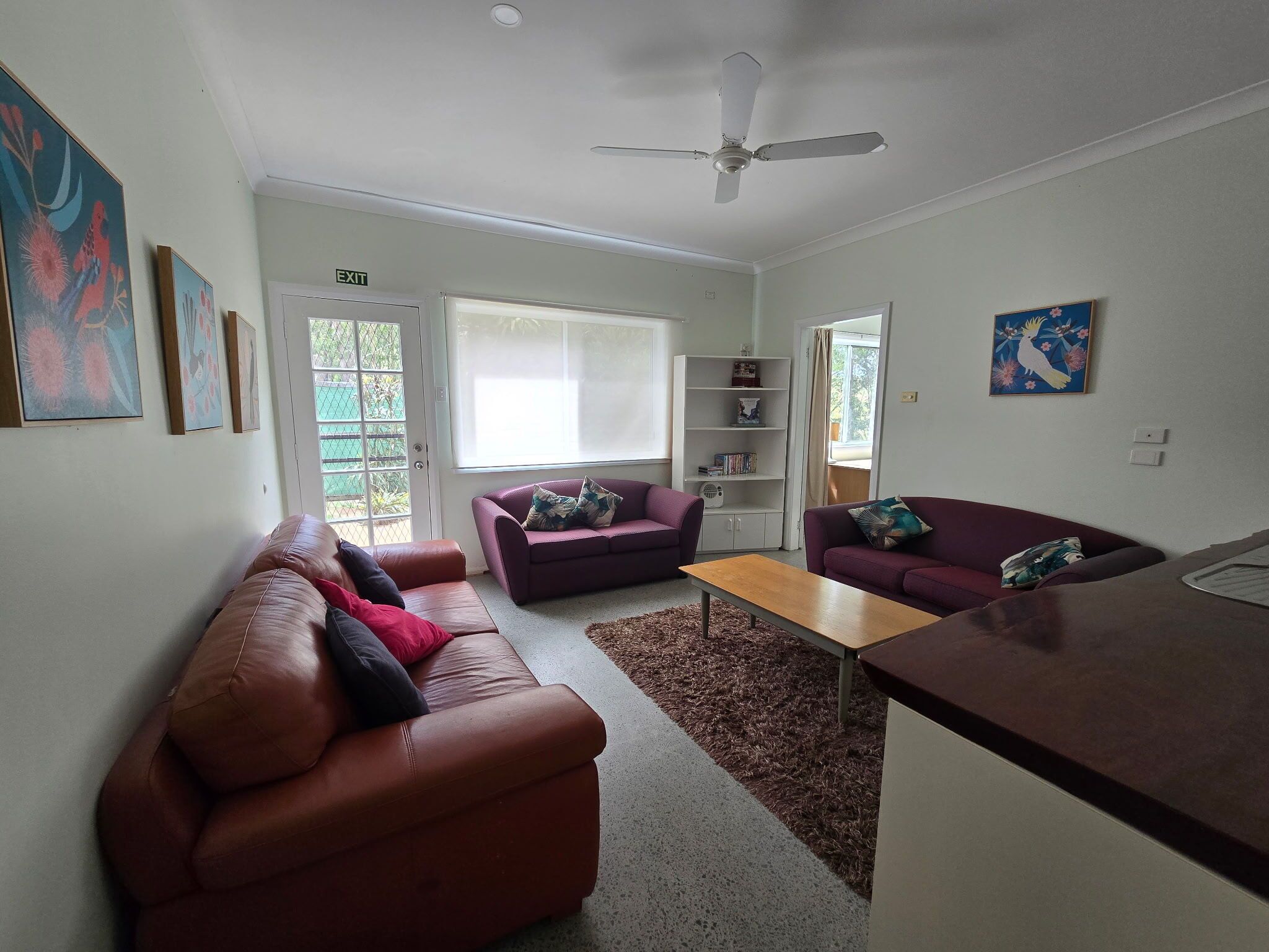 A living room with a brown leather sofa, a purple sofa, a wooden coffee table, and a white bookshelf.