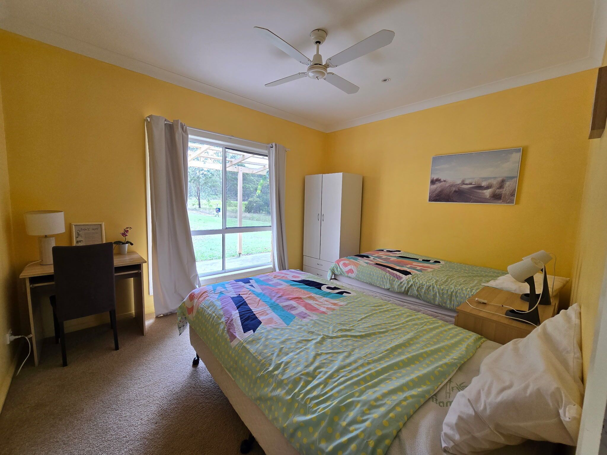 A room with two beds, a desk, a chair, a wardrobe, and a ceiling fan.