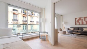 Habitació d'hotel moderna amb una finestra gran, un llit, una taula petita, una cadira, un sofà i una làmpada de peu.