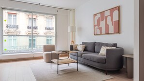 Un saló modern amb un sofà gris, una taula petita, una cadira i una finestra gran que ofereix vistes a un edifici amb balcons.