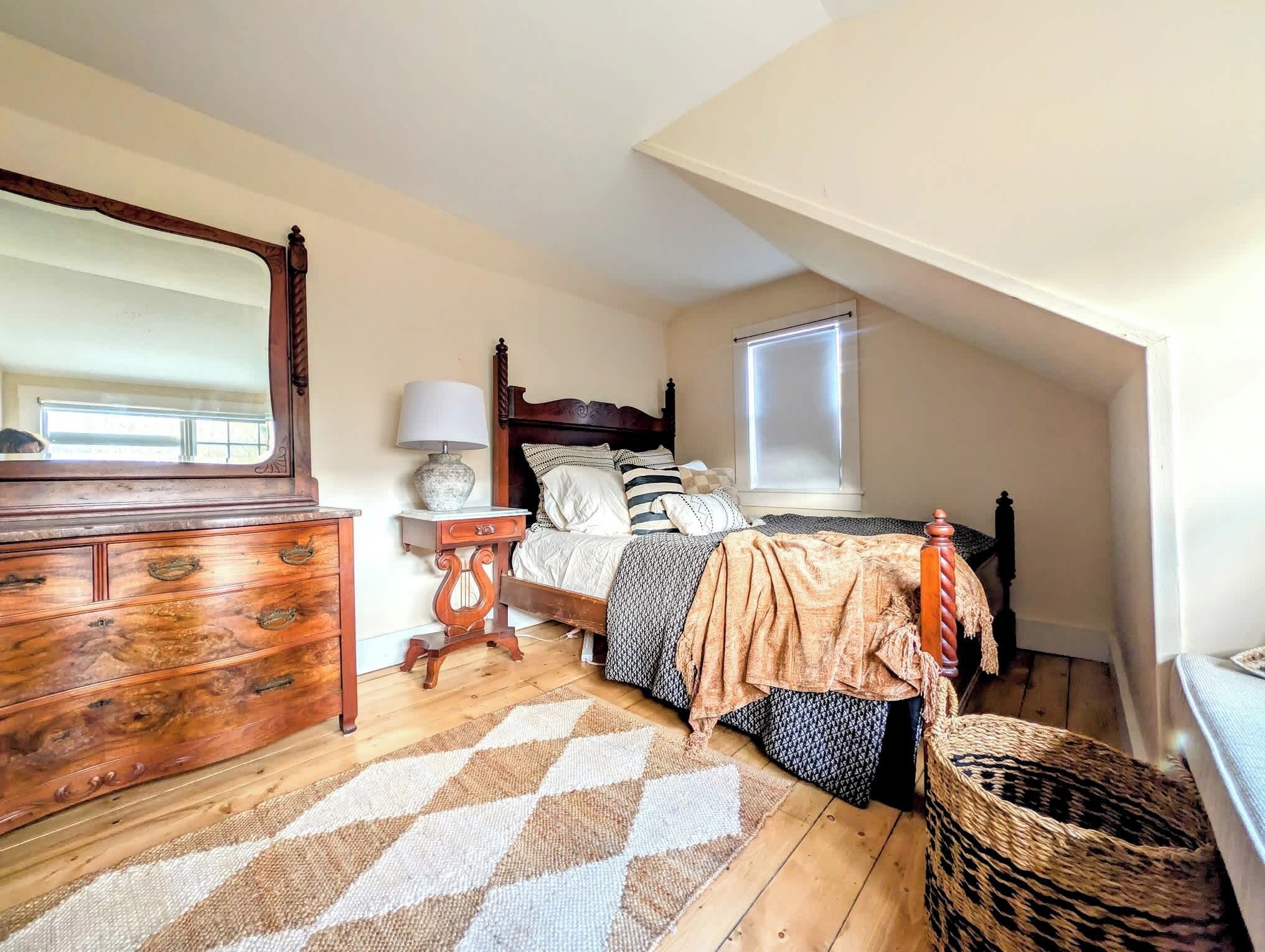 A bedroom with a wooden dresser, a bed with a headboard, a bedside table with a lamp, a window with blinds, and a woven basket.