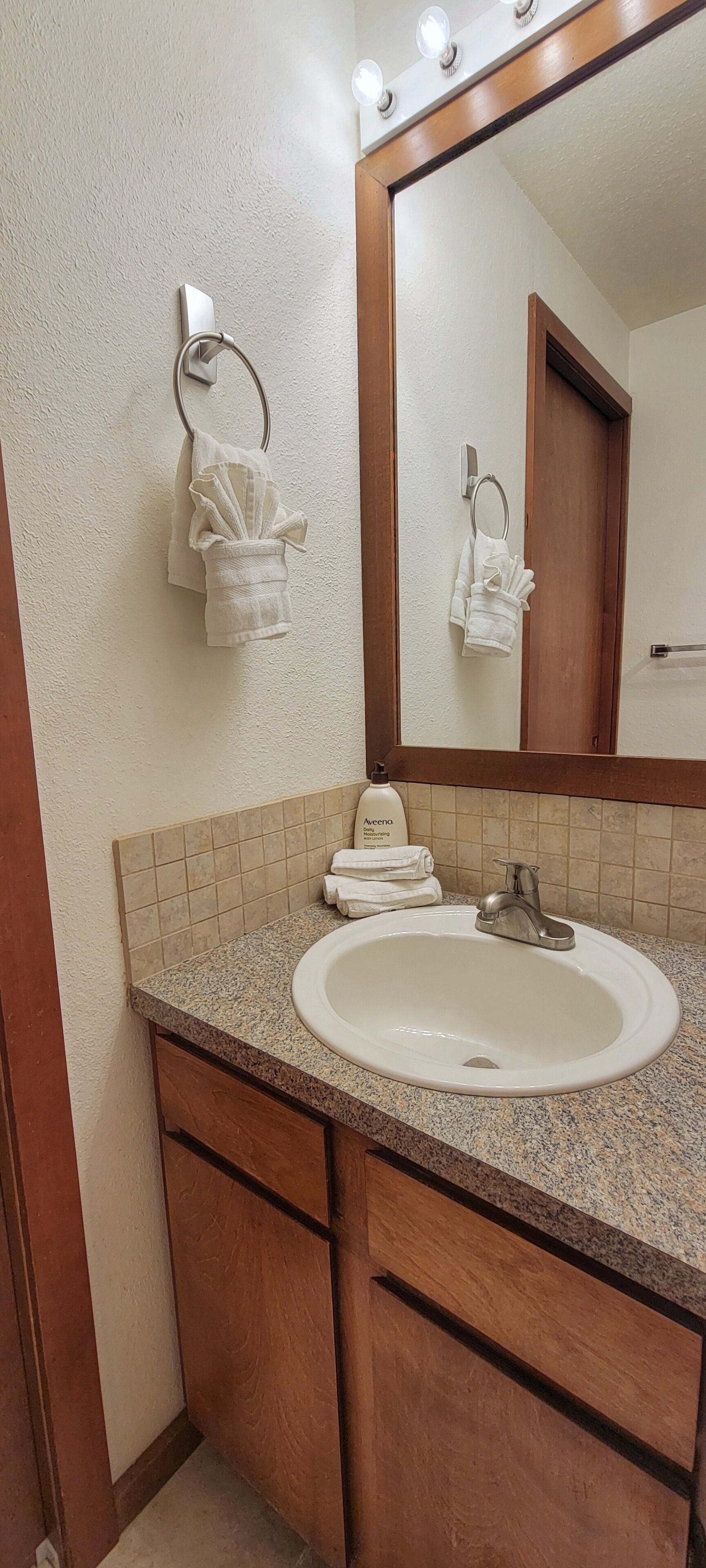 A bathroom with a granite countertop, white sink, and a mirror with lights. There are towels hanging on hooks and a bottle of hand soap on the counter.