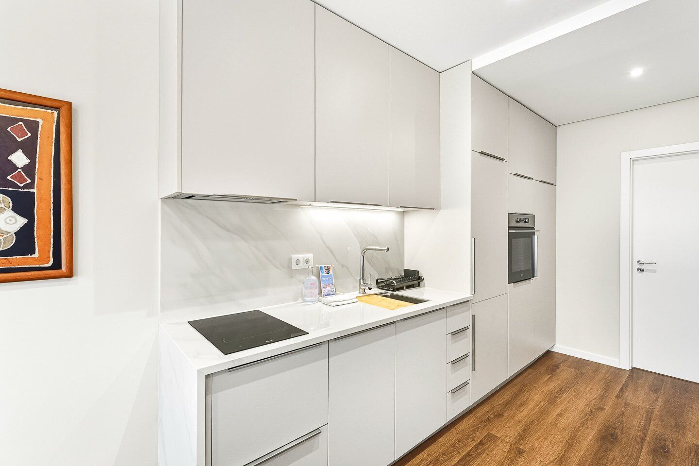 Una cocina moderna con armarios blancos, horno empotrado y encimera con hervidor y tabla de cortar.