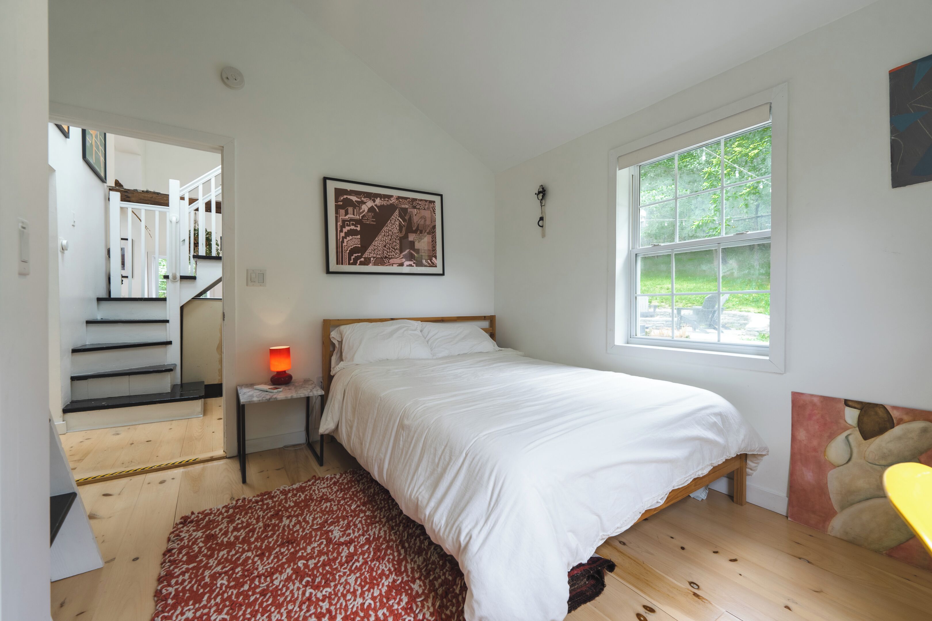 A bedroom with a bed, a nightstand, a red patterned rug, a window with a view of greenery, and a staircase leading to another room.