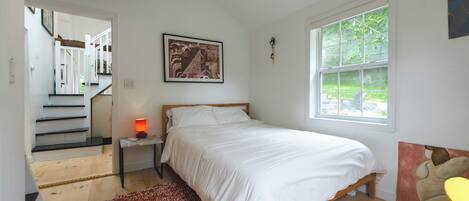 A bedroom with a bed, a nightstand, a red patterned rug, a window with a view of greenery, and a staircase leading to another room.