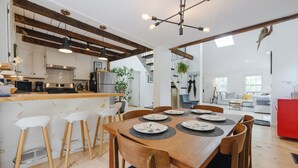 Una cocina moderna con vigas de madera, un comedor con mesa de madera y taburetes de bar, y un luminoso salón con un armario rojo.
