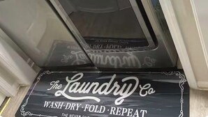 Máquina de lavar com carga frontal, painel de controle e uma placa com a inscrição 'The Laundry Co Wash-Dry-Fold-Repeat'.
