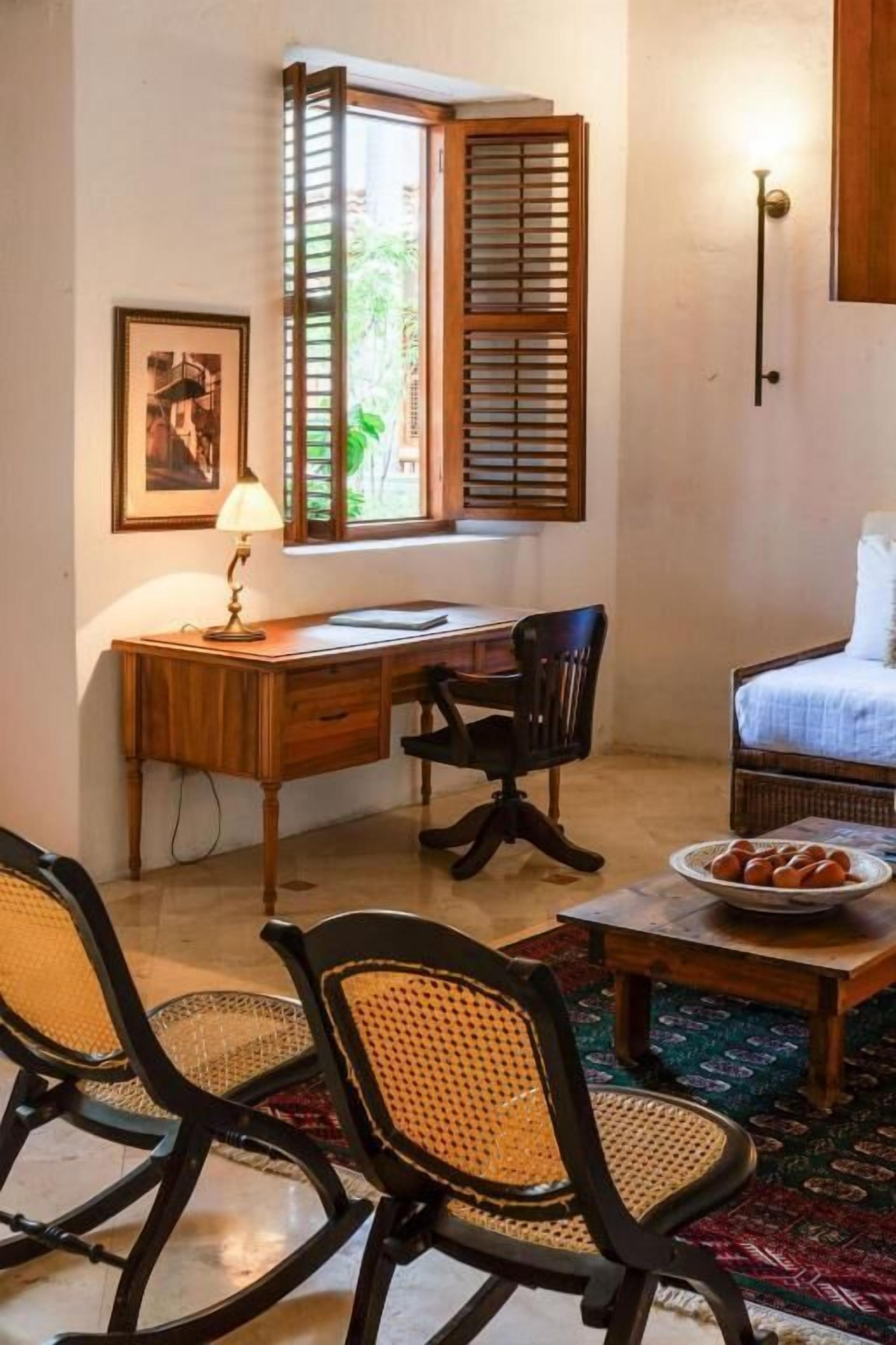 Una habitación compacta con escritorio de madera, dos sillas, cama, ventana con contraventanas de madera, lámpara de pared e imagen enmarco.