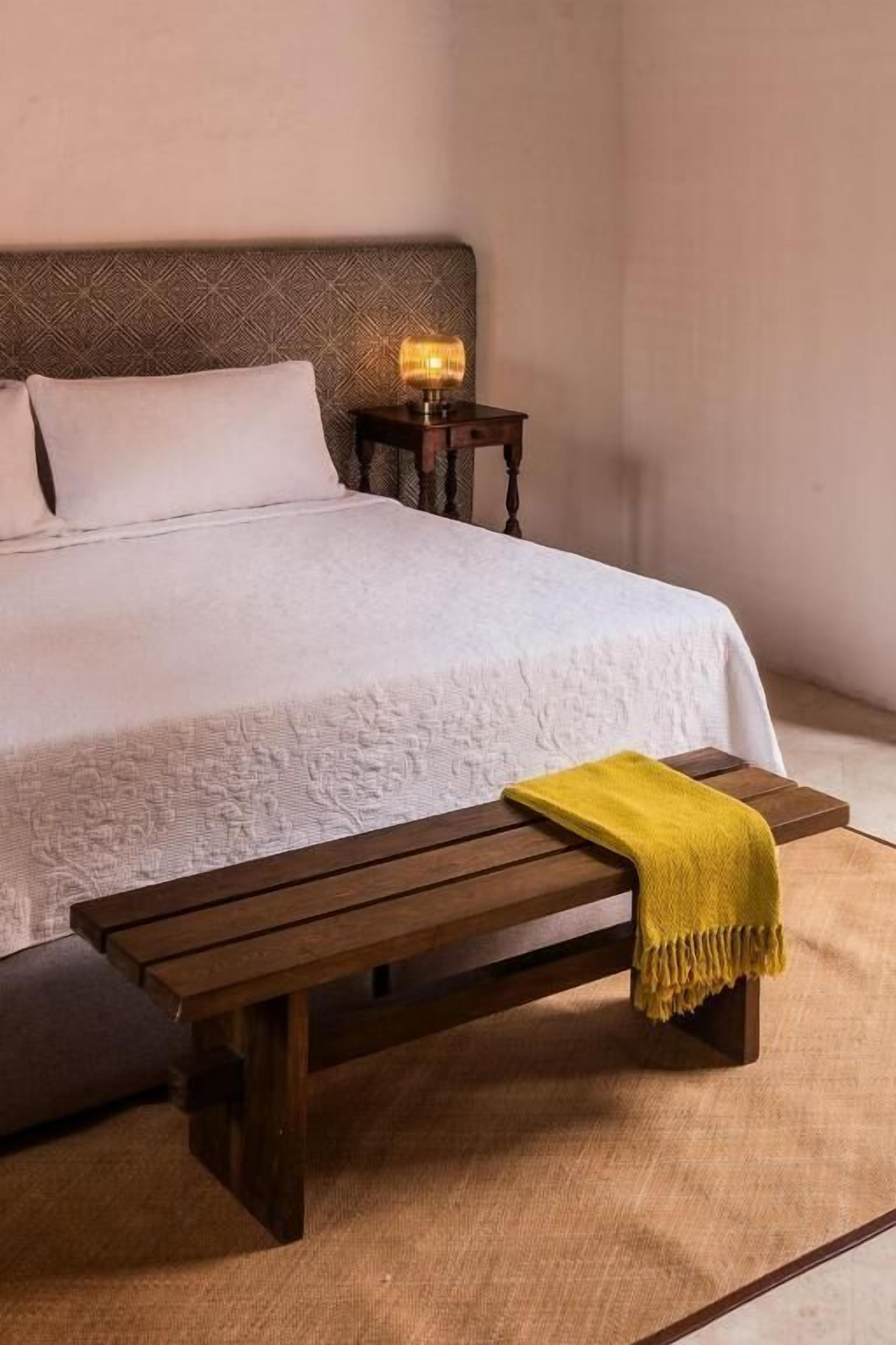 A neatly made bed with a white duvet, a wooden bench with a yellow throw, and a nightstand with a lamp.