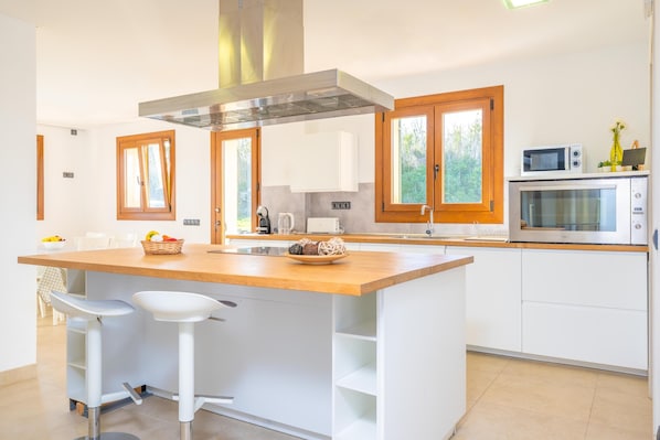 Una cocina moderna con una isla central, armarios blancos, detalles de madera y ventanas amplias.