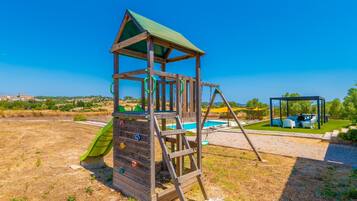A wooden play structure with a slide and climbing holds, a swing set, and a pool area with outdoor seating.