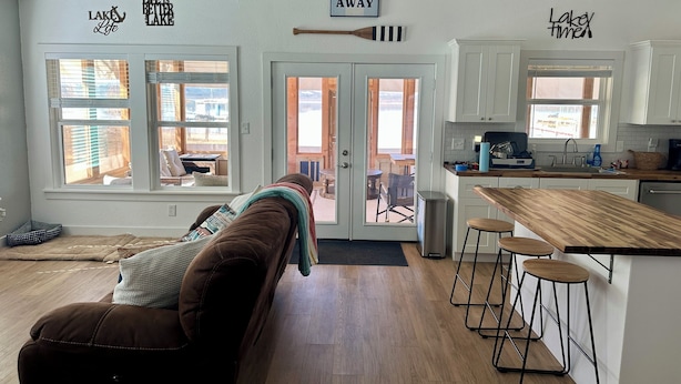 A cozy living room with a brown couch, a dining area with a wooden table and bar stools, and a kitchen with white cabinets and a wooden countertop. - Lakefront Home with Private Beach & Dock, Great Sunsets, 7 Miles from Rocker B (Graford)