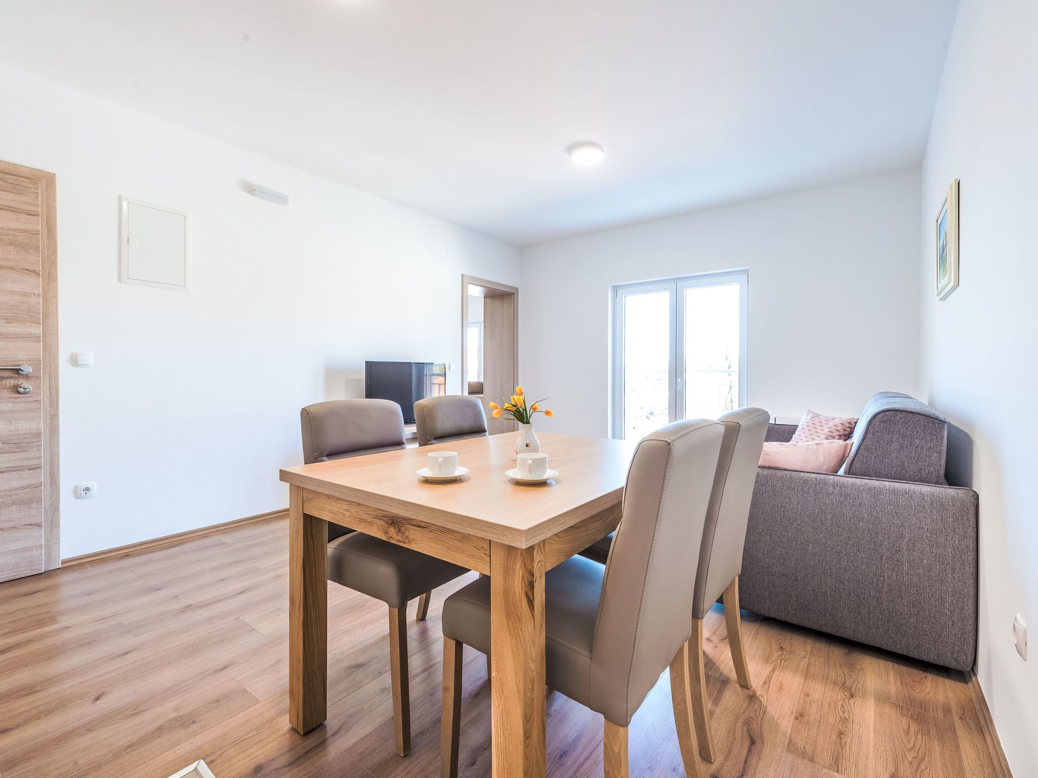 A modern living room with a wooden dining table, two chairs, a grey sofa, and a window with natural light.