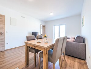 A modern living room with a wooden dining table, two chairs, a grey sofa, and a window with natural light. - Wohnungen Rilovic - Wohnung A3 by Interhome (Dubrovnik/Cavtat)