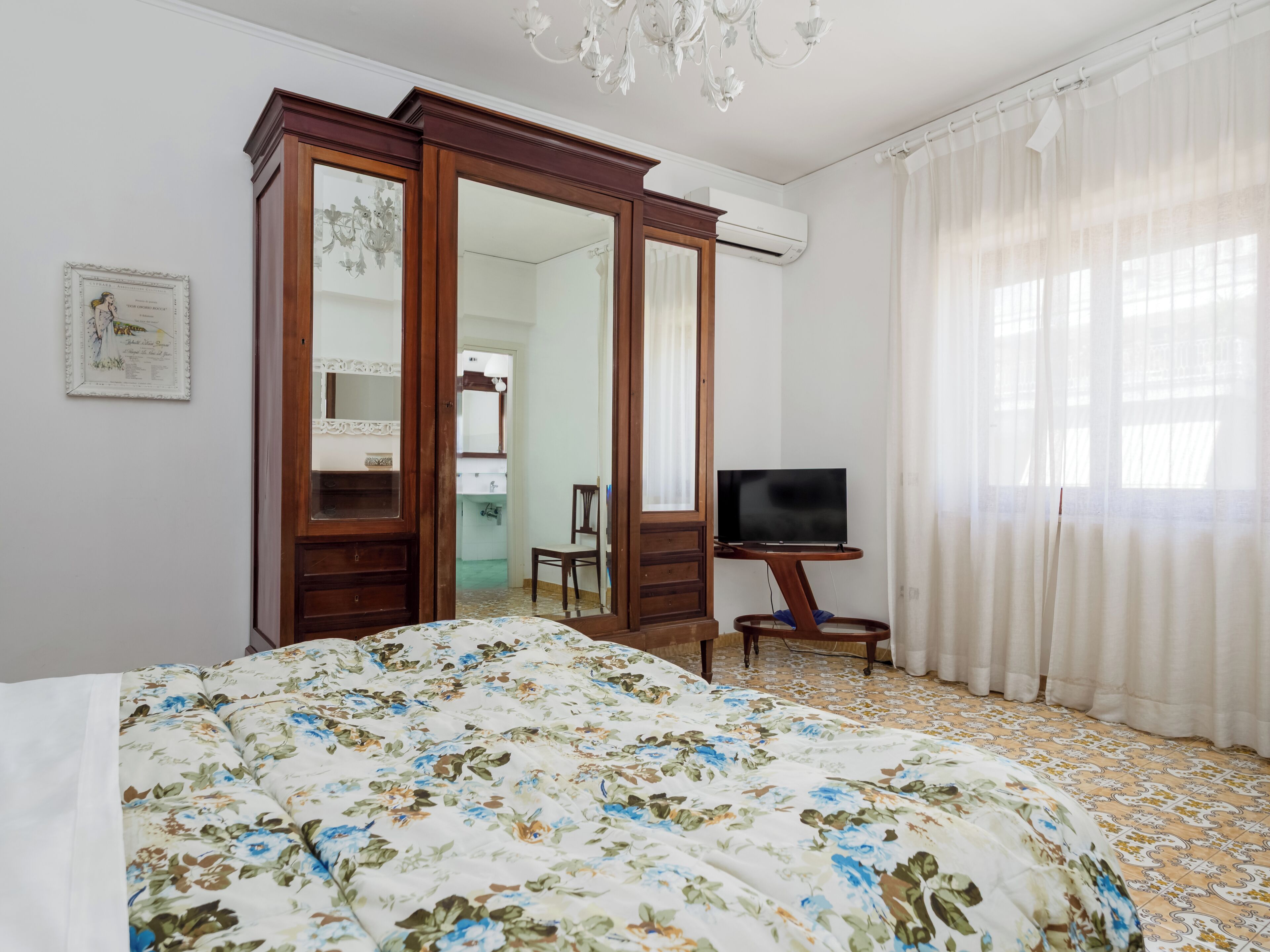 A bedroom with a bed, a TV on a stand, a wardrobe, and a bathroom visible through an open door.