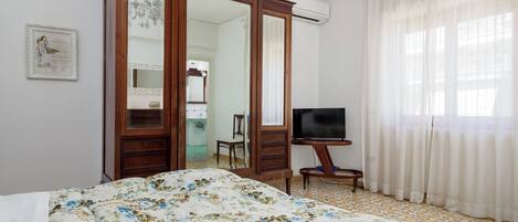 A bedroom with a bed, a TV on a stand, a wardrobe, and a bathroom visible through an open door.