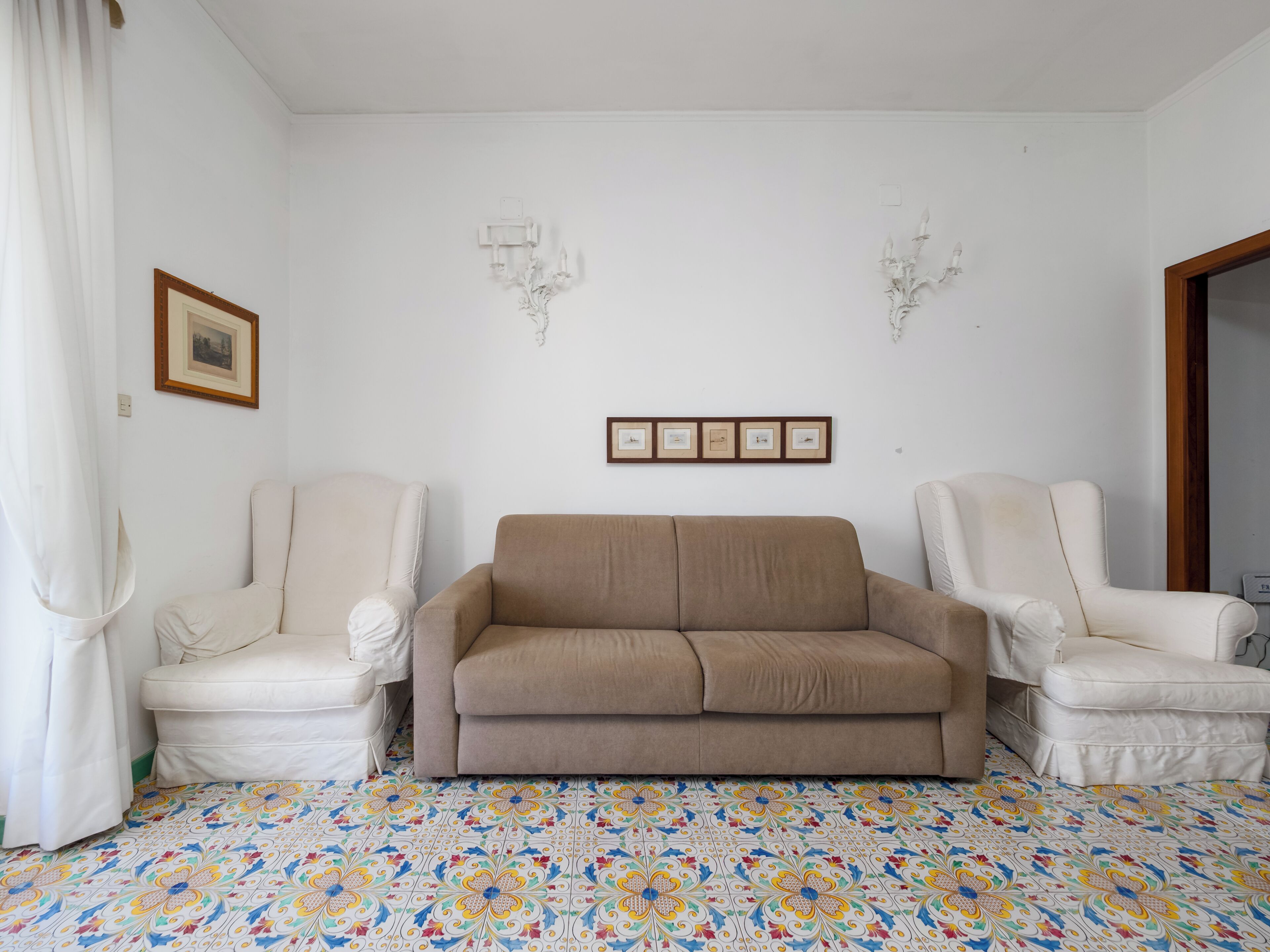 A room with a beige sofa, two white armchairs, a patterned floor, and wall-mounted picture frames.