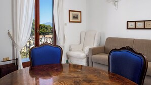 Ein Zimmer mit Esstisch, zwei Stühlen, einer Couch, einem Fenster mit Vorhängen, einem gerahmten Bild und einer Wandleuchte.