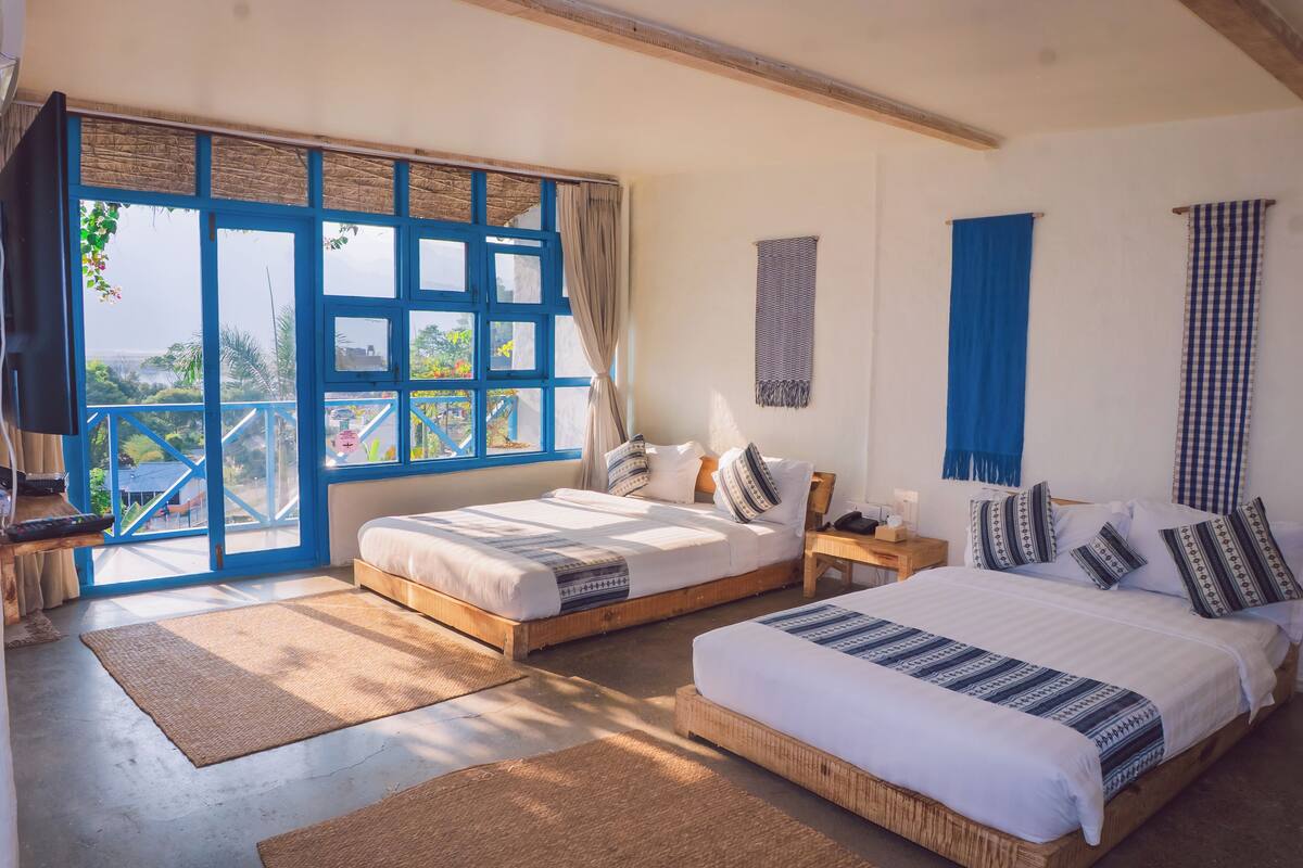 A room with two beds, a large window with blue shutters, and a view of greenery outside.