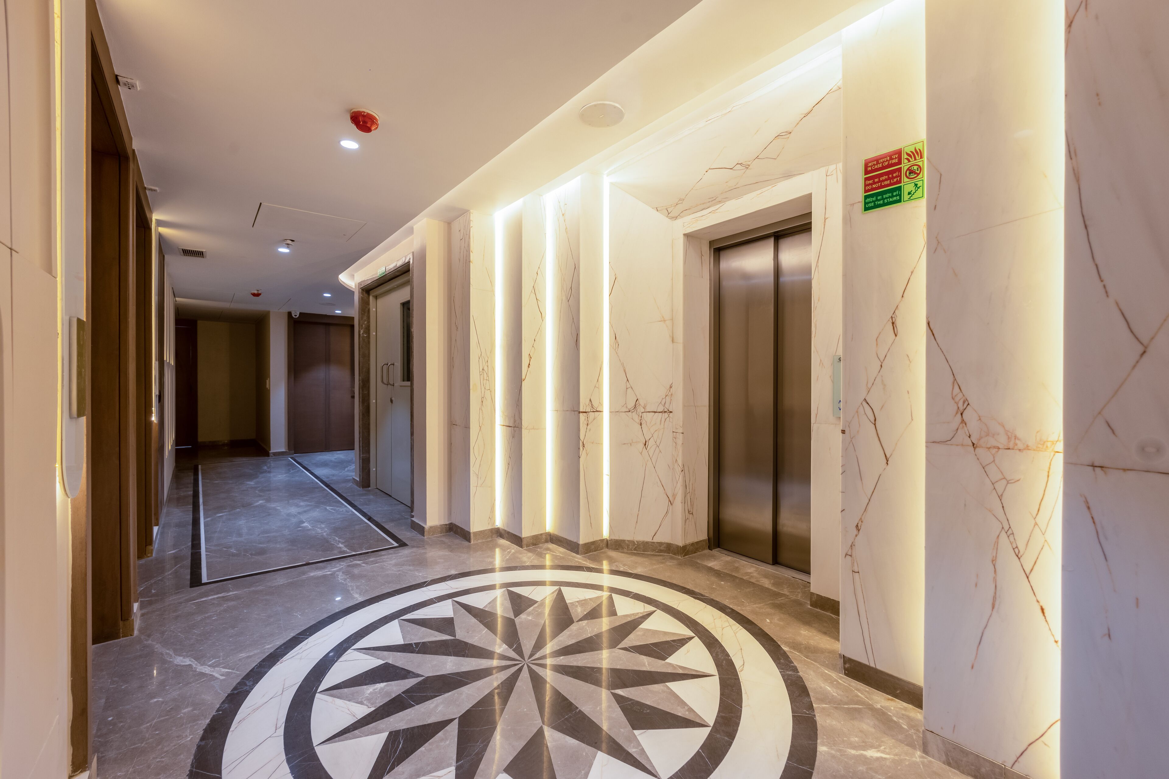 Een moderne hotelgang met marmeren vloeren, een geometrisch patroon en een rookmelder aan het plafond.
