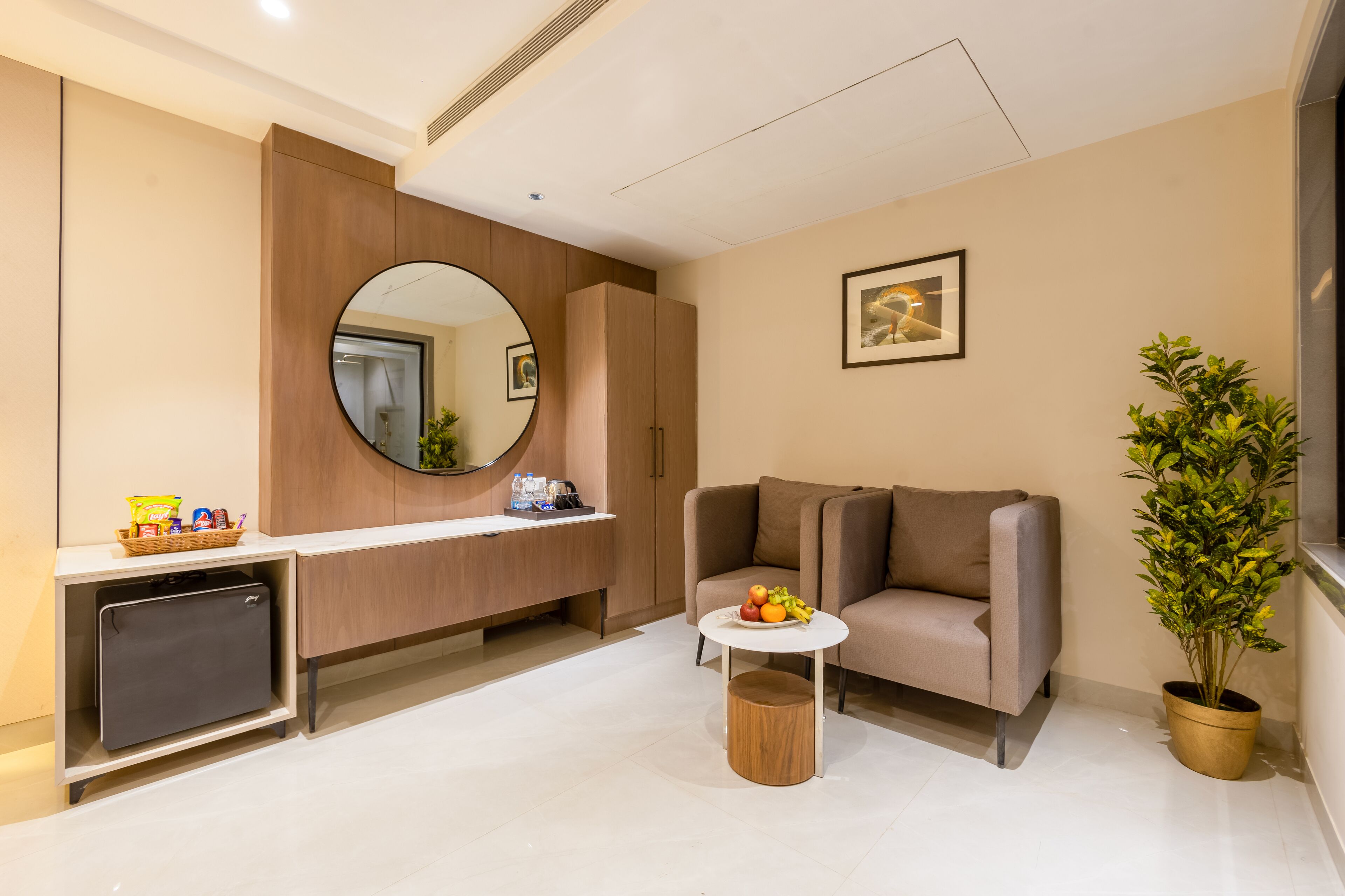 Een moderne hotelkamer met een bank, een klein rond tafeltje met fruit, een compacte koelkast, een grote spiegel en een potplant.