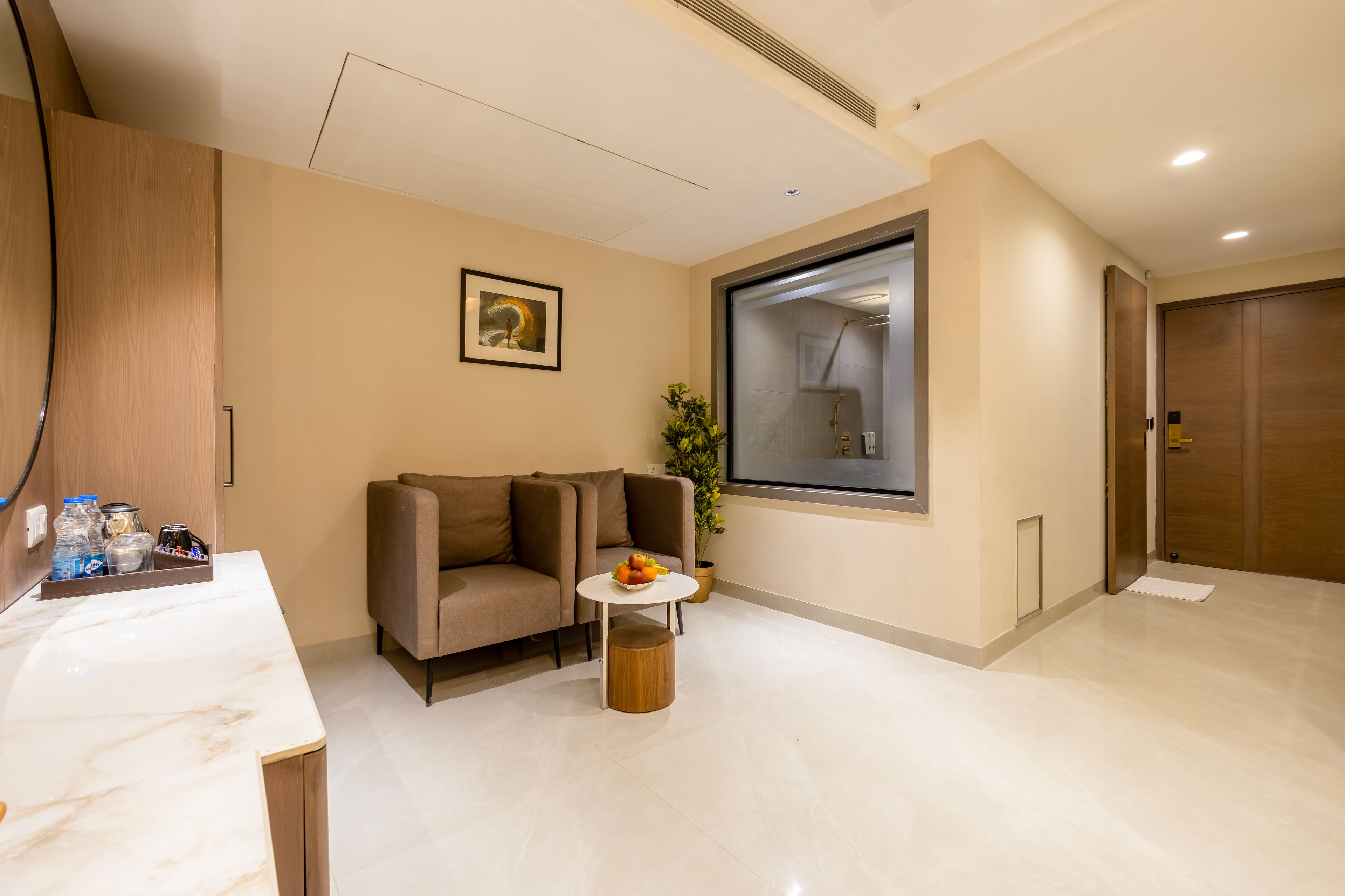 Ein modernes Hotelzimmer mit Marmorplatte, einer Couch, einem kleinen runden Tisch mit Früchten, einer Topfpflanze und einem großen Fenster mit Dusche.