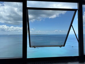 A view through a window with a metal frame, overlooking a large body of water and distant land. - Apartment in the Salvador Carnival circuit - sea view and beautifully decorated. (Salvador)