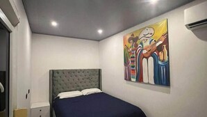 Un dormitorio con cama, mesita de noche, aire acondicionado de pared y un cuadro de colores.
