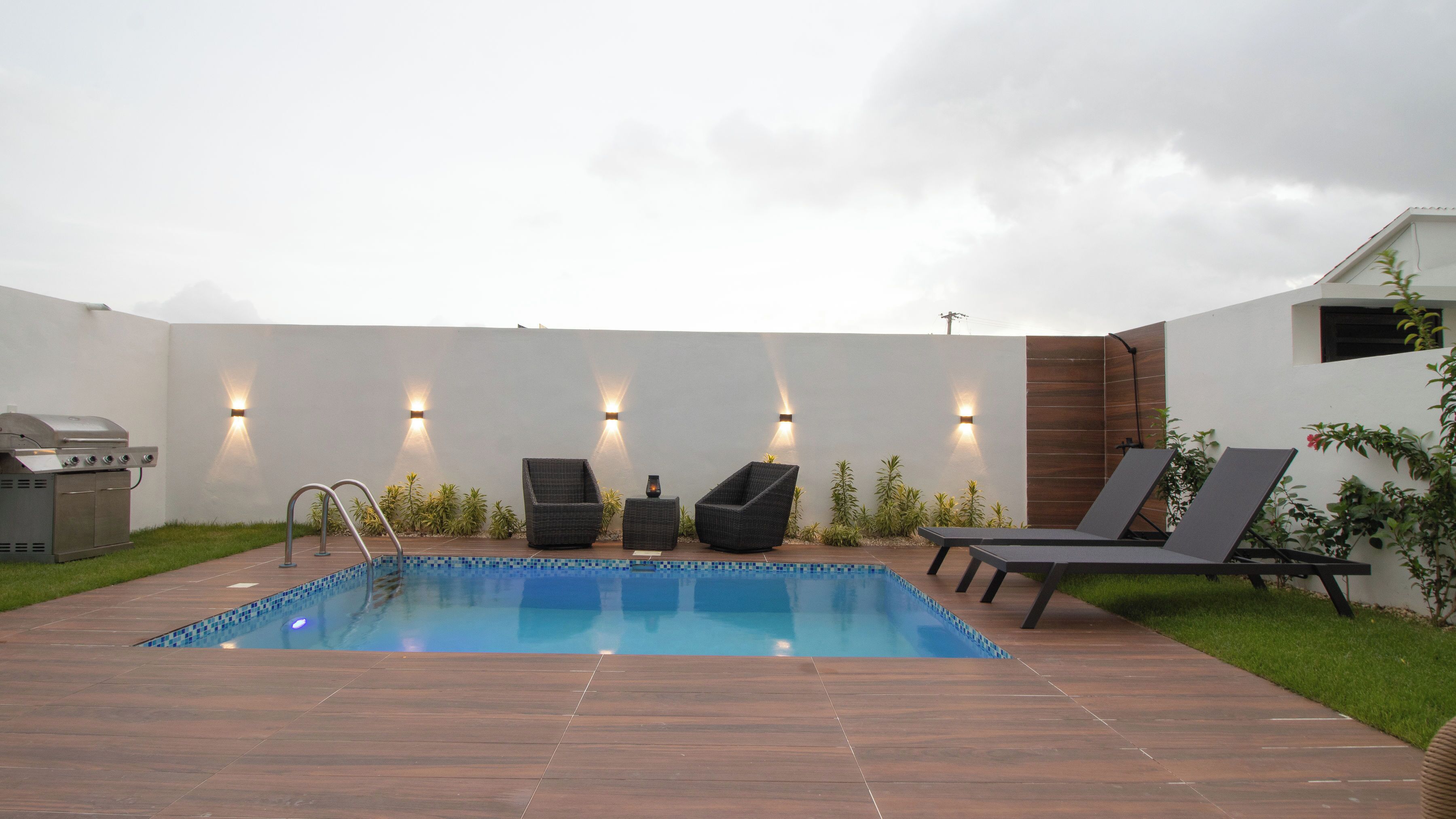 A swimming pool with a wooden deck, two lounge chairs, a barbecue grill, and wall-mounted lights.