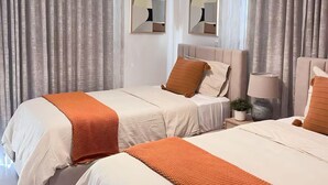 Ett modernt hotellrum med två sängar, ett stort fönster med tunna gardiner, en hängande lampa och ett litet nattduksbord med en lampa och en växt.