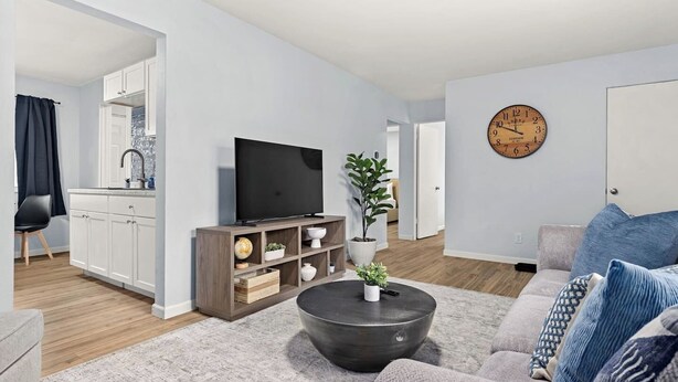 A modern living room with a flat-screen TV, a coffee table, and a sofa. There is a kitchen area with cabinets and a sink visible in the background. - Home Suite Home: E-Minutes to Downtown & P Heights (Peoria)