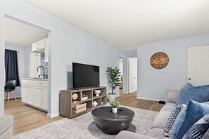A modern living room with a flat-screen TV, a coffee table, and a sofa. There is a kitchen area with cabinets and a sink visible in the background.