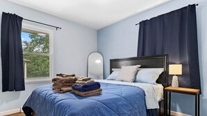 Un dormitorio con una cama, una mesita de noche con lámpara, una ventana con cortinas y una pila de toallas.