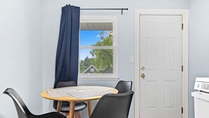Una habitación pequeña con una mesa y dos sillas, una ventana que da a una casa, y una puerta blanca.