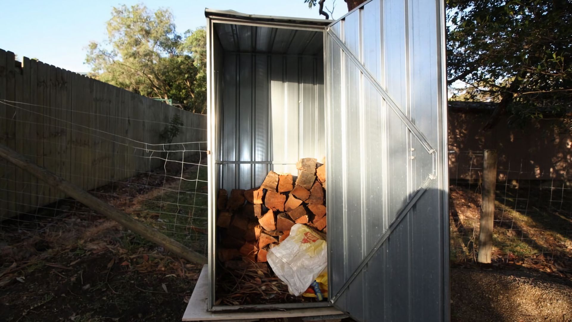 A metal shed with a door open, revealing a stack of firewood and a plastic bag inside.