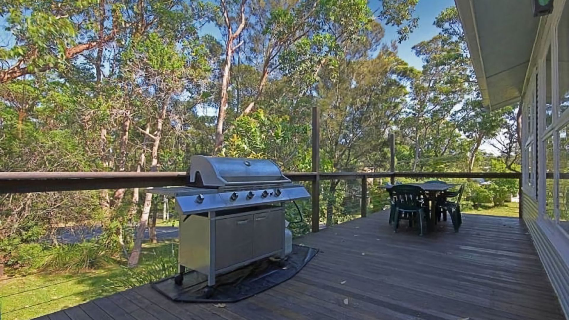 A covered outdoor deck with a barbecue grill, a table with chairs, and a view of trees.