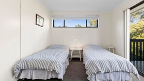 A room with two single beds, a small bedside table with a lamp, a window with a view of trees, and a balcony with a railing. - Halliwag Haven is perfect for a family getaway (Cunjurong Point)