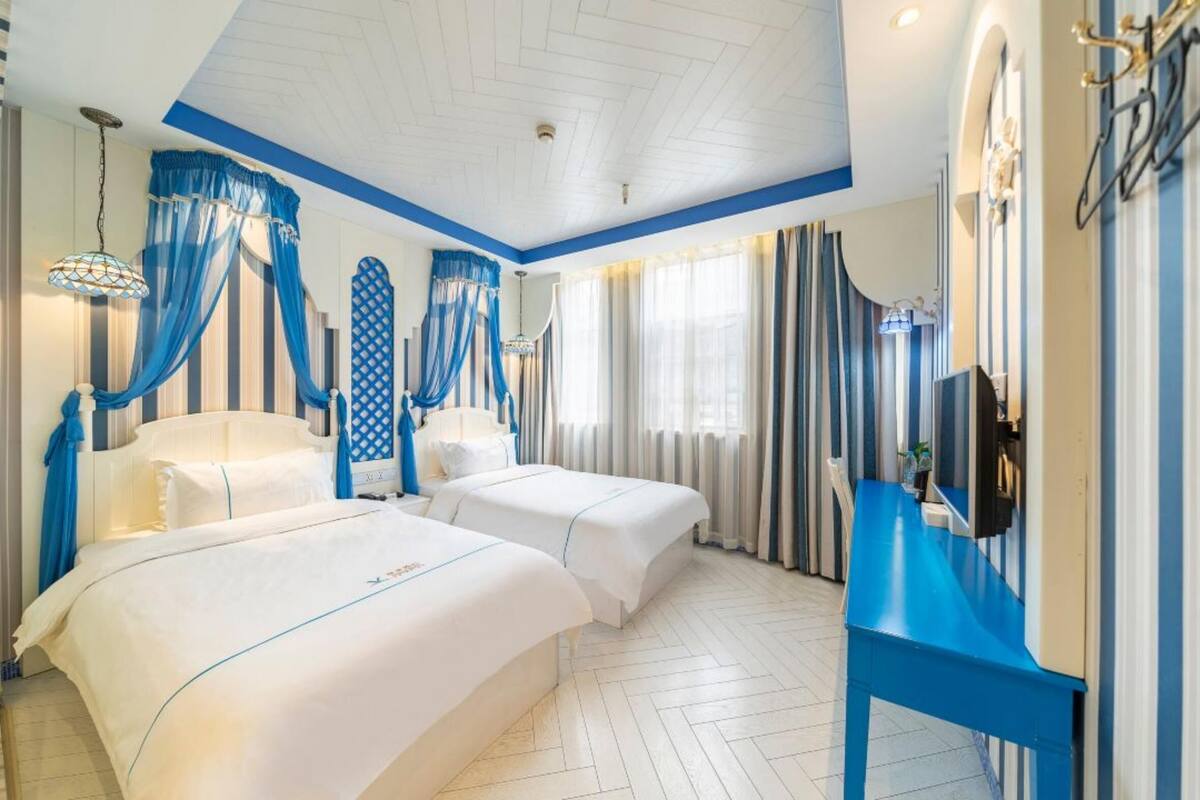 Ein Hotelzimmer mit zwei Betten, einer blauen Akzentwand und einem großen Fenster mit Vorhängen.