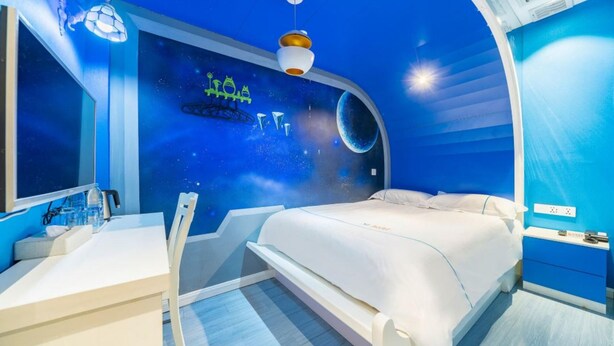 Une chambre à coucher avec un lit, un bureau et une chaise. Un des murs de la pièce est bleu et décoré d’une fresque sur le thème de l’espace.