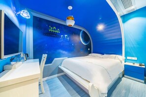 Une chambre à coucher avec un lit, un bureau et une chaise. Un des murs de la pièce est bleu et décoré d’une fresque sur le thème de l’espace.