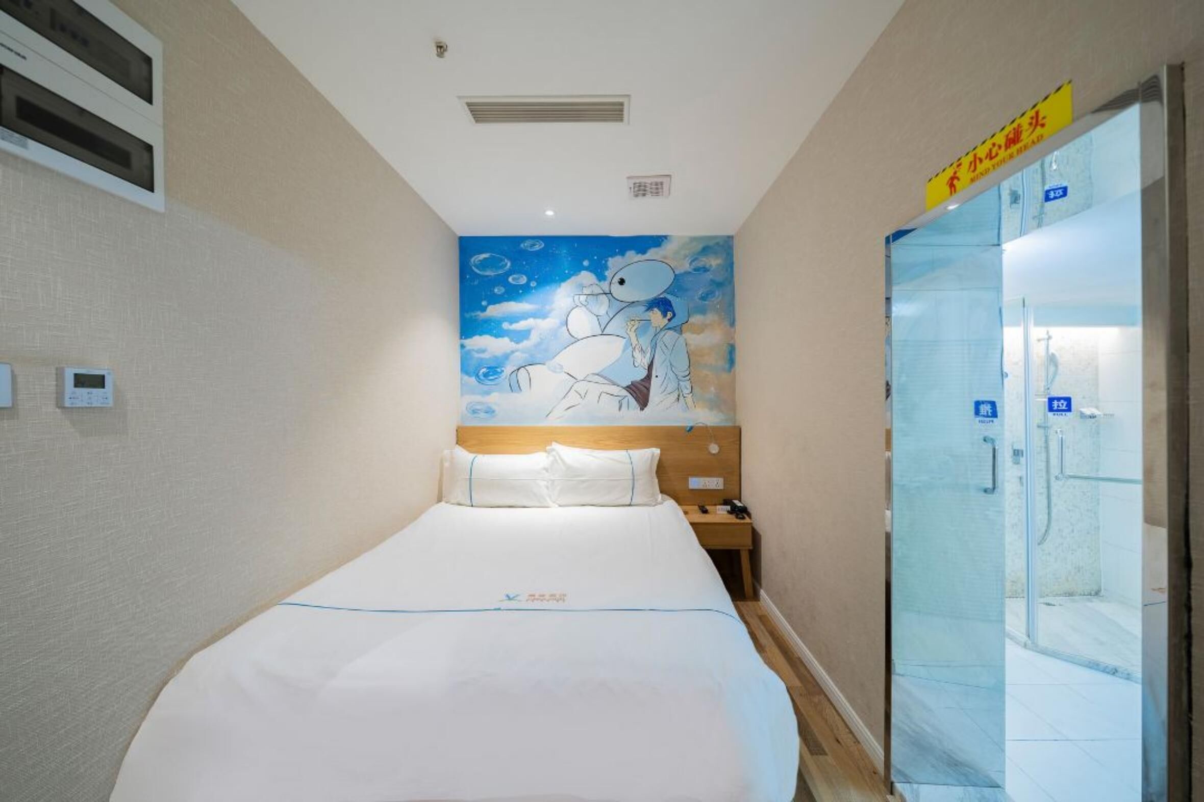 A hotel room with a bed, a nightstand, a wall mural, a bathroom with glass doors, and a sign above the door.