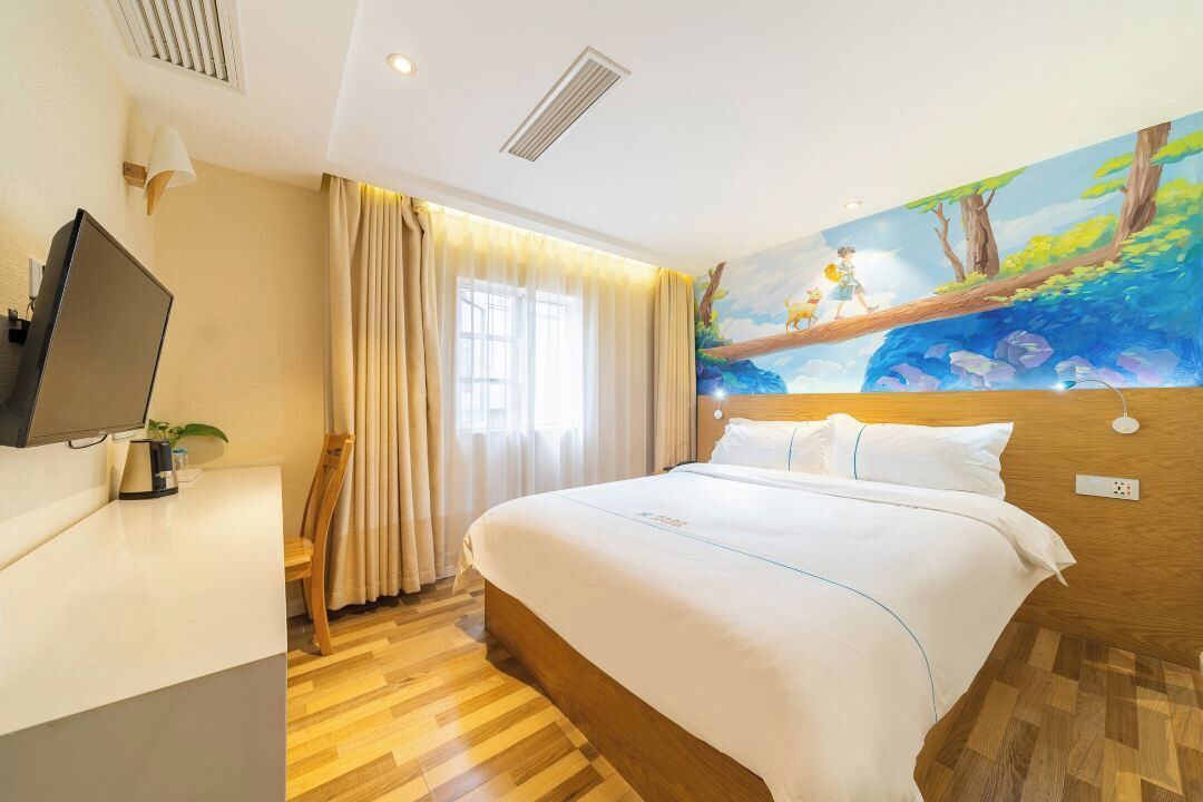 A hotel room with a large bed, a flat-screen TV, a desk with a lamp, and a mural of a forest scene on the wall.