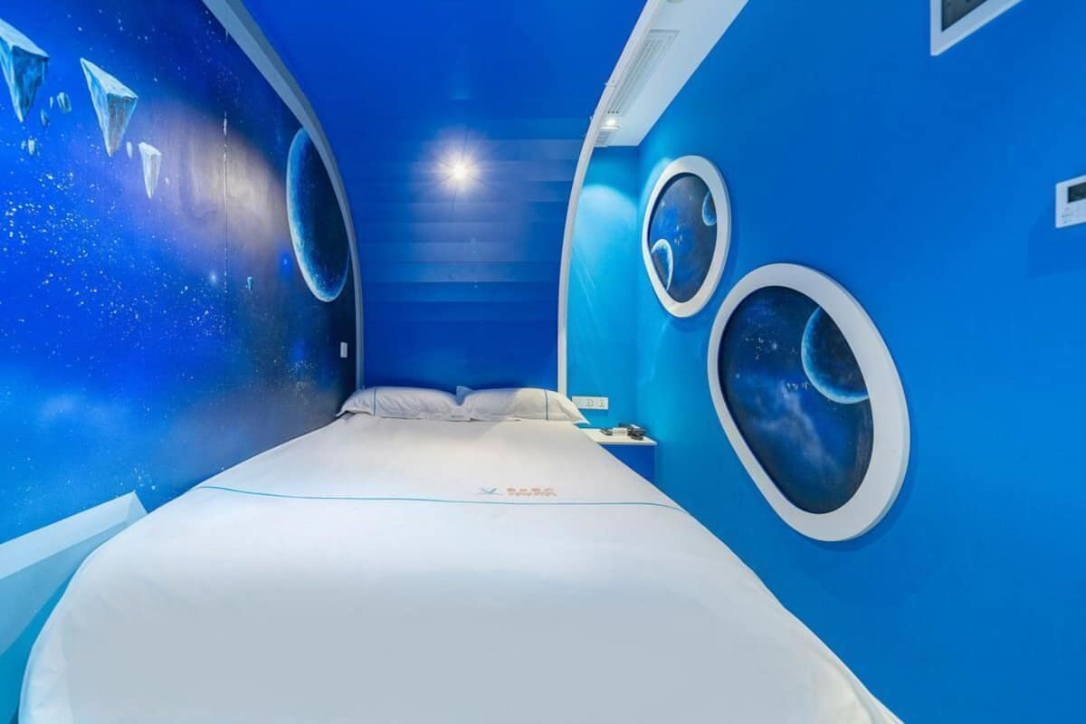 Ein Zimmer mit einem Bett, einem Nachttisch und zwei runden Fenstern mit Wandgemälden zum Thema Weltall.